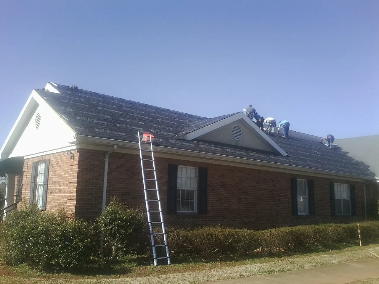Crew tearing shingles off a brick ranch roof with a ladder set — Emergency Roof Repair work by our crew in Romeo