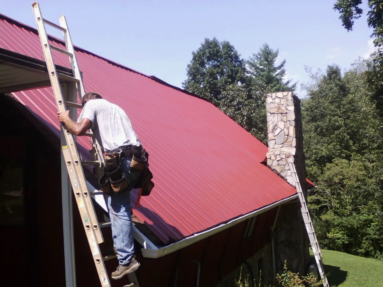 Roofer on a ladder beside a red metal roof and stone chimney — Soffit & Fascia Repair by Roofing Brothers in Romeo
